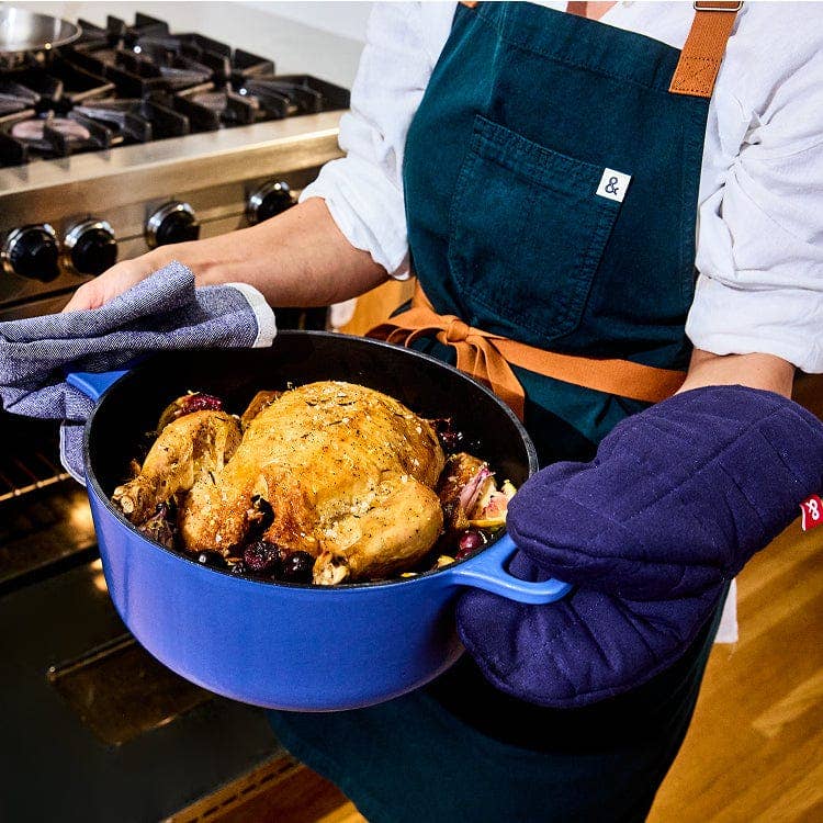 Person holding a blue cast iron skillet with a roasted chicken, wearing an apron and oven mitts.