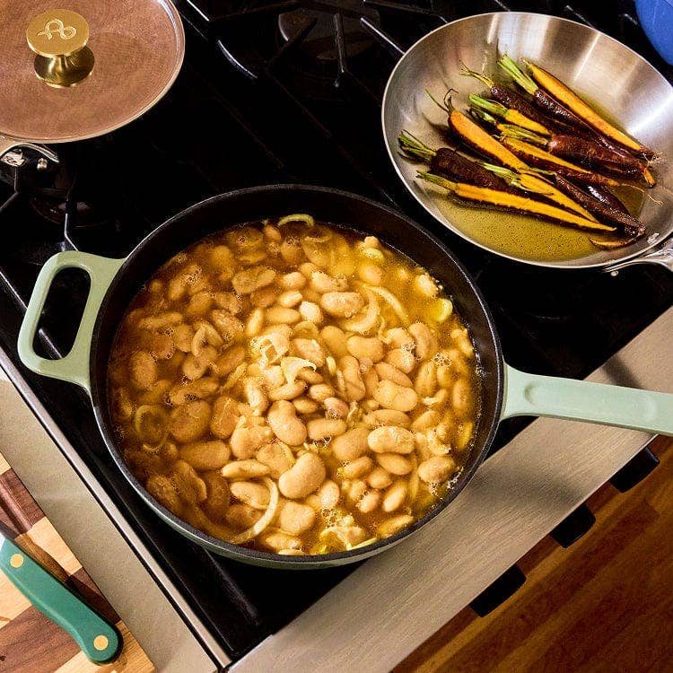 A black cast iron skillet with a sage green handle sits on a black stovetop, filled with a bean and onion stew. A stainless steel pan with carrots and red chilies is visible in the upper right, and a wooden cutting board with a green-handled knife is in the lower left.