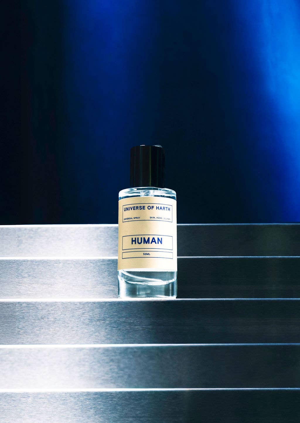 A bottle of UNIVERSE OF MANKIND fragrance named HUMAN, featuring a cream-colored label and a black cap, placed on a metallic silver staircase against a dark blue background.