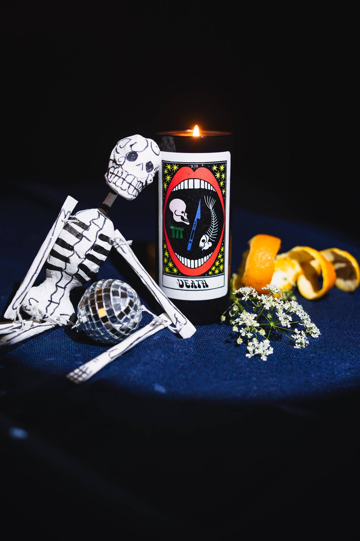 A black jar candle with a Day of the Dead 'Death' tarot card design, lit and standing next to a white skeleton figurine, a small disco ball, orange peels, and white flowers on a dark blue surface against a black background.