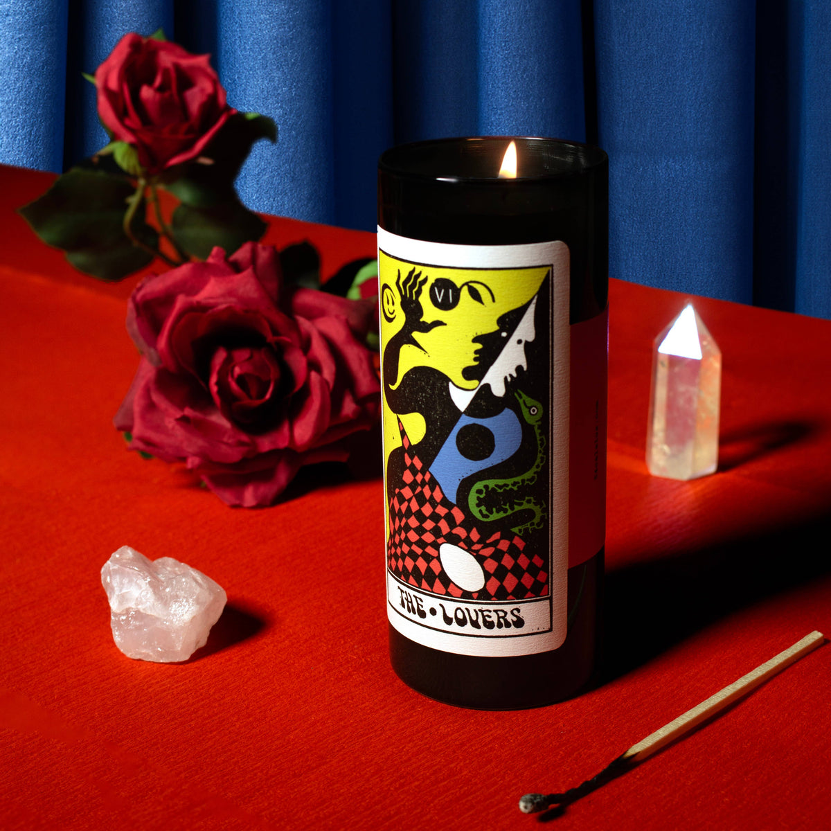 Candle labeled 'The Lovers' with roses and crystals on a red surface against a blue curtain.