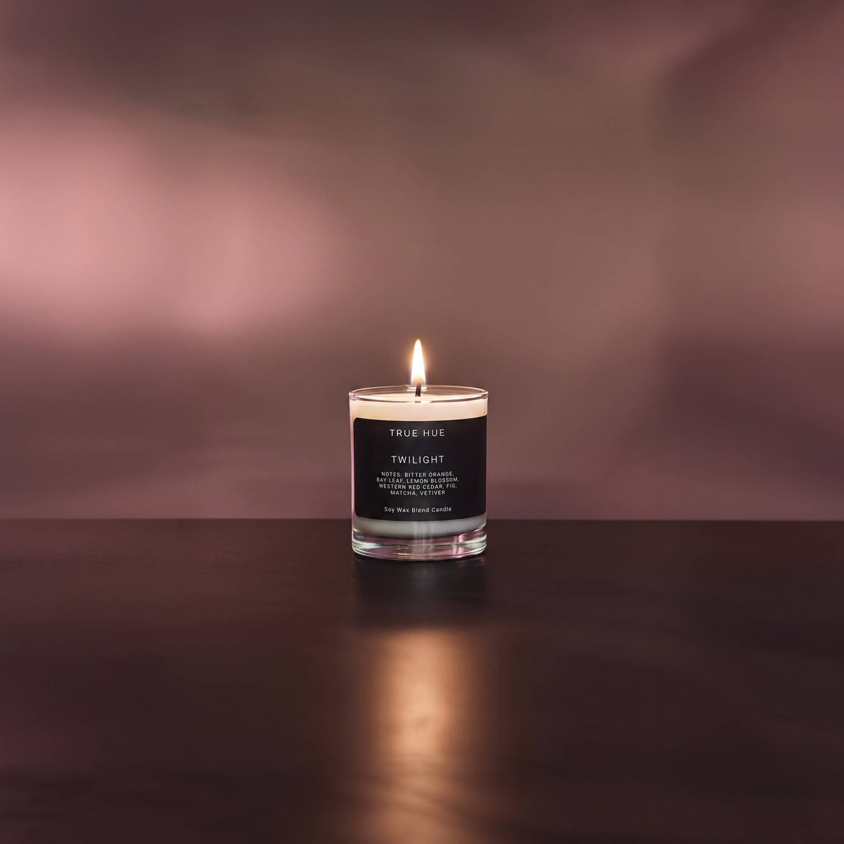 A lit candle in a clear glass jar with a black label reading 'TRUE MUE TWILIGHT', placed on a dark reflective surface against a soft pinkish-brown blurred background.