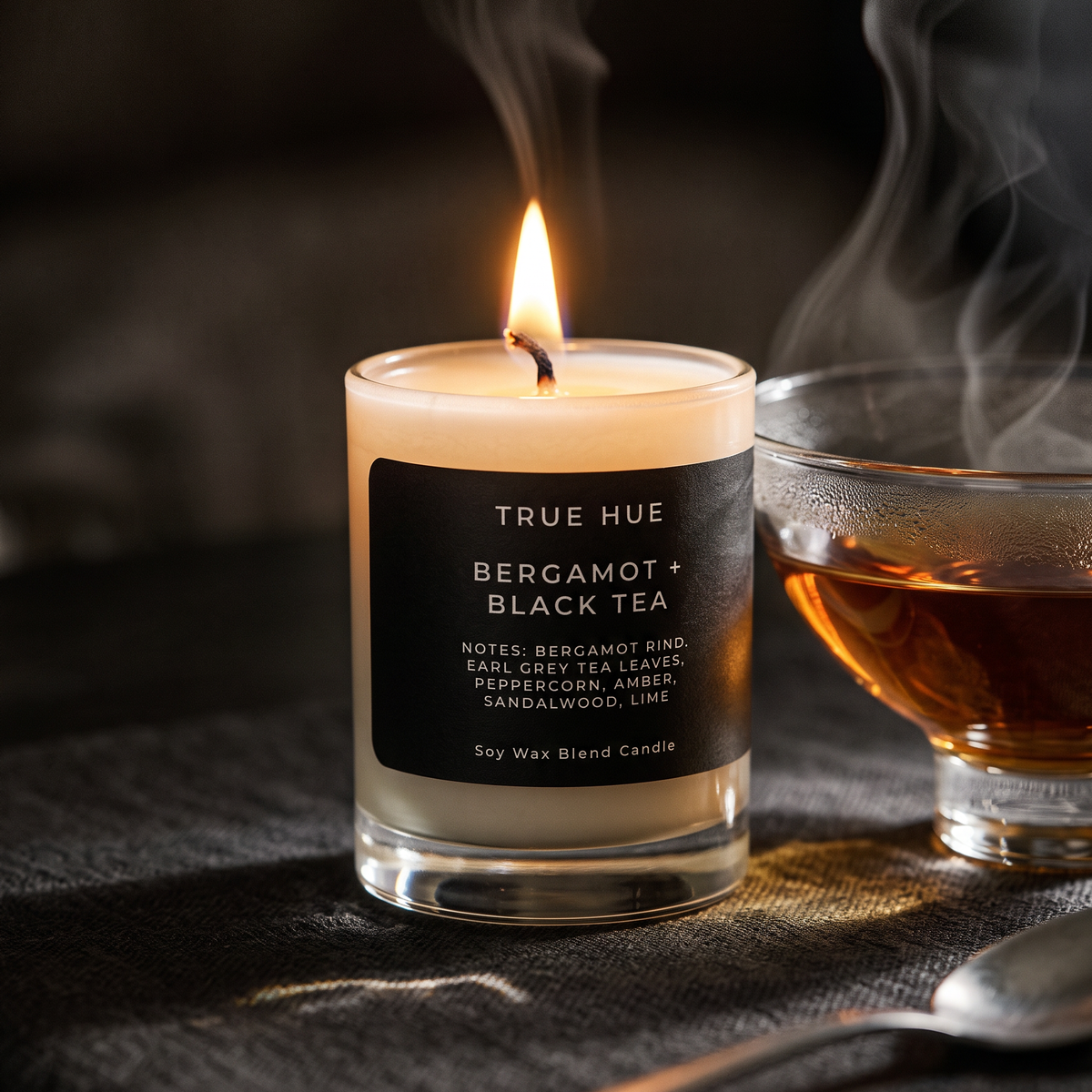 A lit soy wax candle in a clear glass jar with a black label reading TRUE HUE BERGAMOT + BLACK TEA, set on a dark surface beside a glass of amber tea with visible steam.