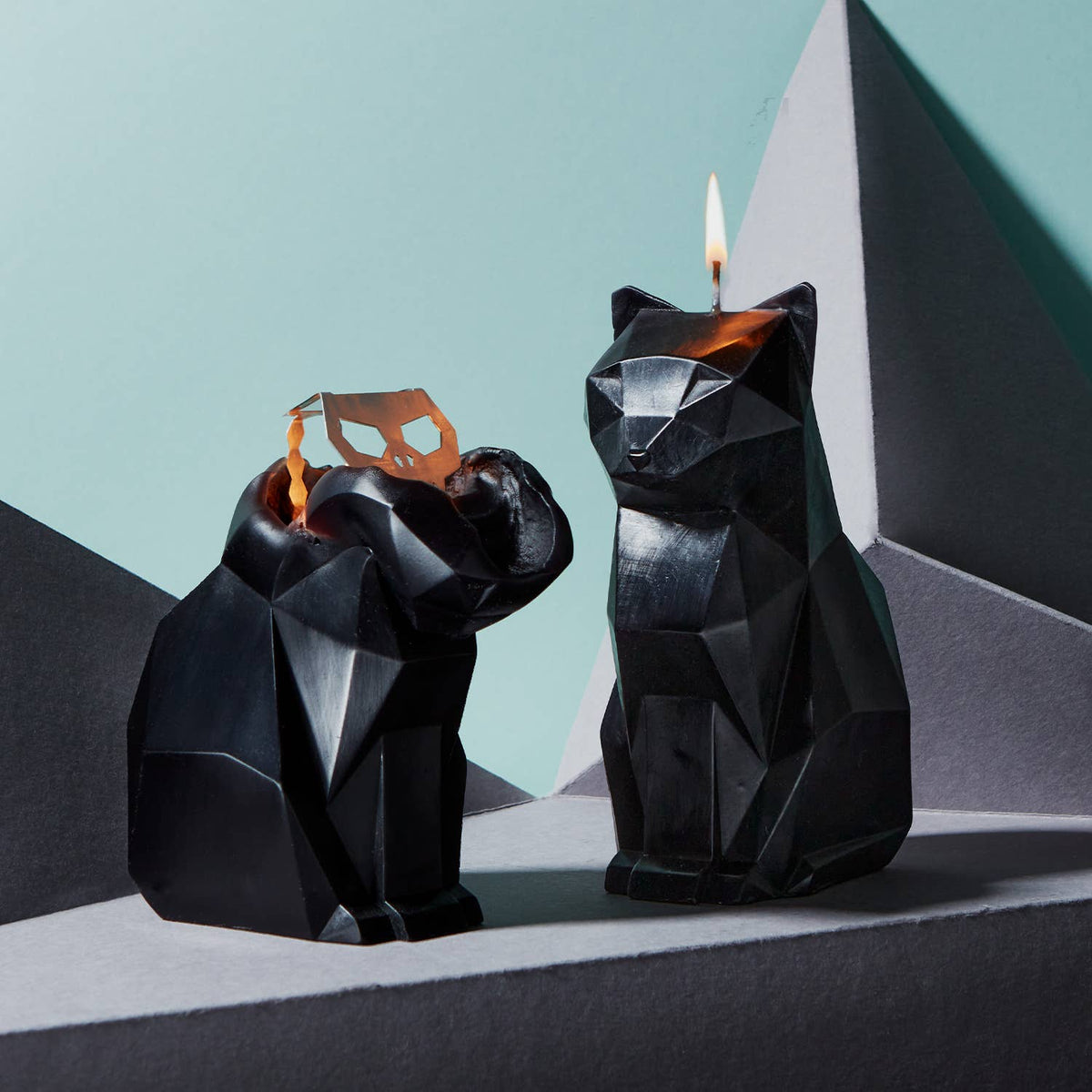 Two black geometric cat-shaped candles with lit wicks against a light blue background