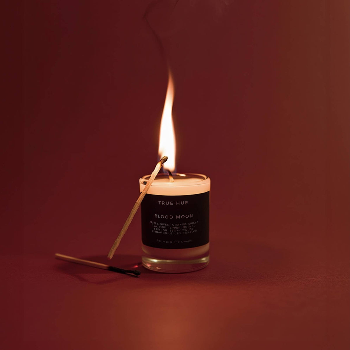 A lit candle in a clear glass jar with a black label reading TRUE HUE and BLOOD MOON, shown on a dark warm red-brown background with a match leaning against the jar and another match on the surface.