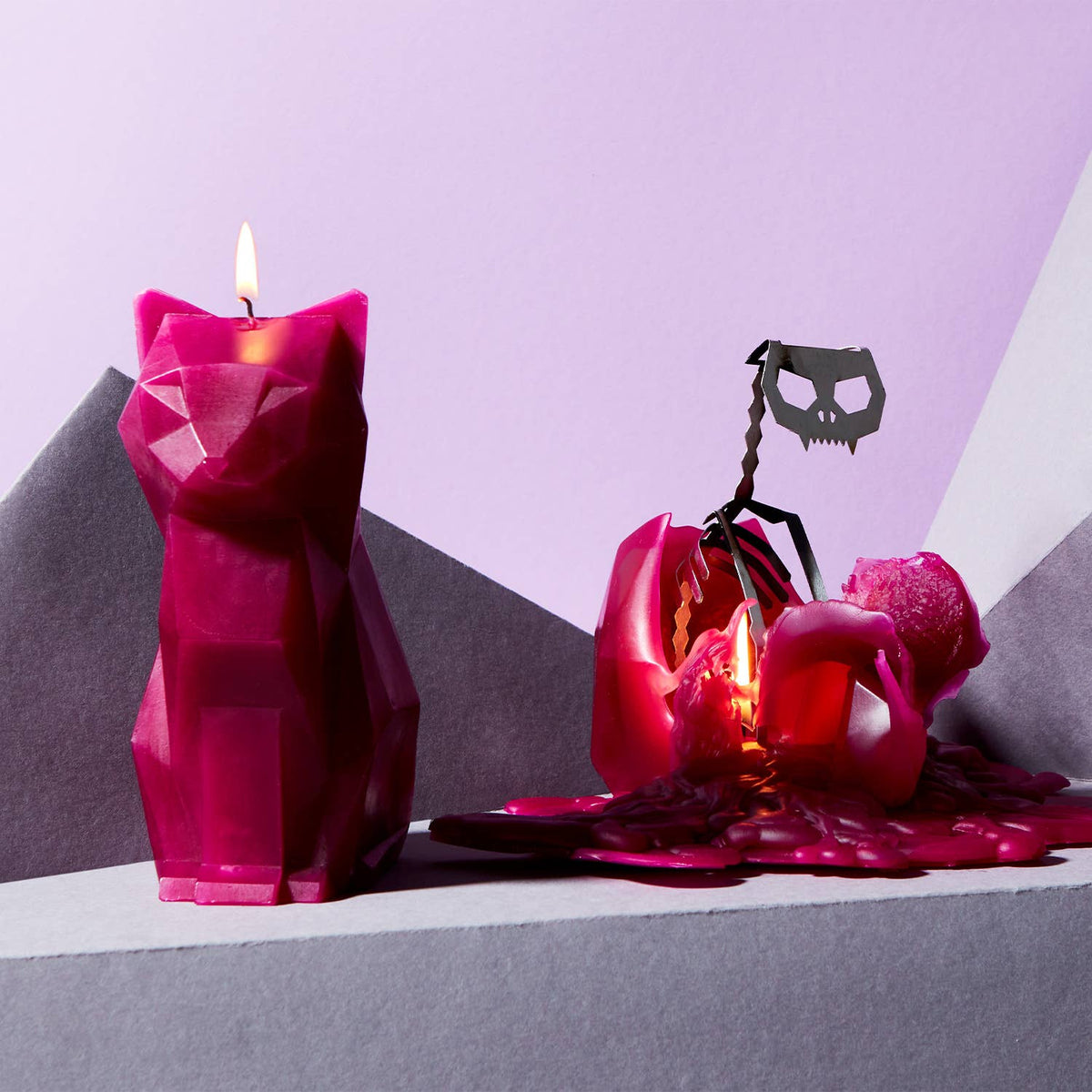 Pink geometric candle shaped like a cat with a skull decoration on a gray and purple background.