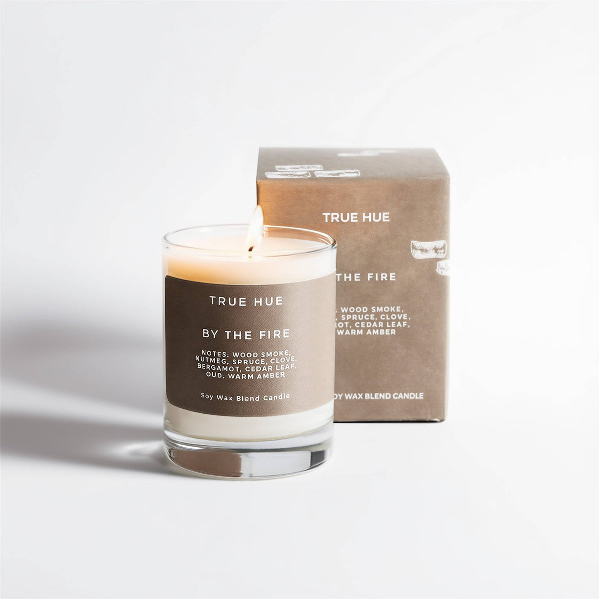 A lit TRUE HUE BY THE FIRE soy wax blend candle in a clear glass jar with a brown label, standing in front of its matching brown cardboard box on a clean white background.