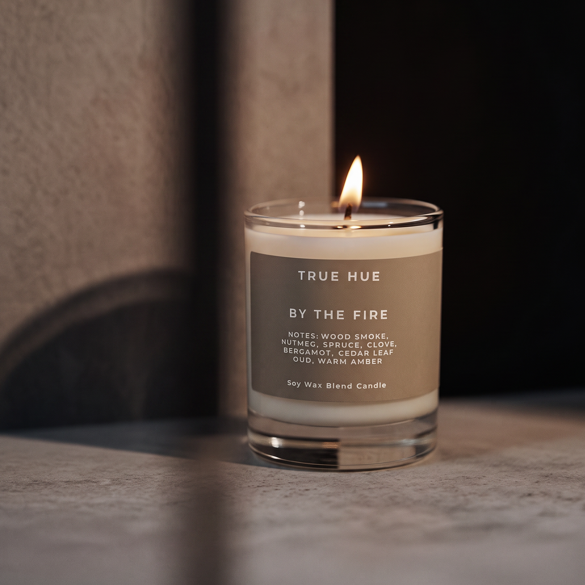 A lit True Hue by the Fire scented soy candle in a clear glass jar, set on a stone-like surface with a warm, dimly lit background.