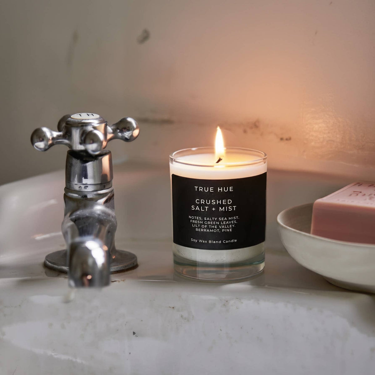 A lit glass jar candle labeled TRUE HUE CRUSHED SALT + MIST sits on a bathroom sink next to a chrome faucet and a pink soap bar in a bowl.