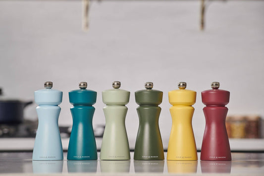 Set of six colorful pepper mills on a reflective surface with a blurred kitchen background.
