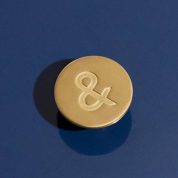 A round gold-toned button or pin featuring an embossed ampersand symbol, photographed on a dark blue background with a soft shadow.