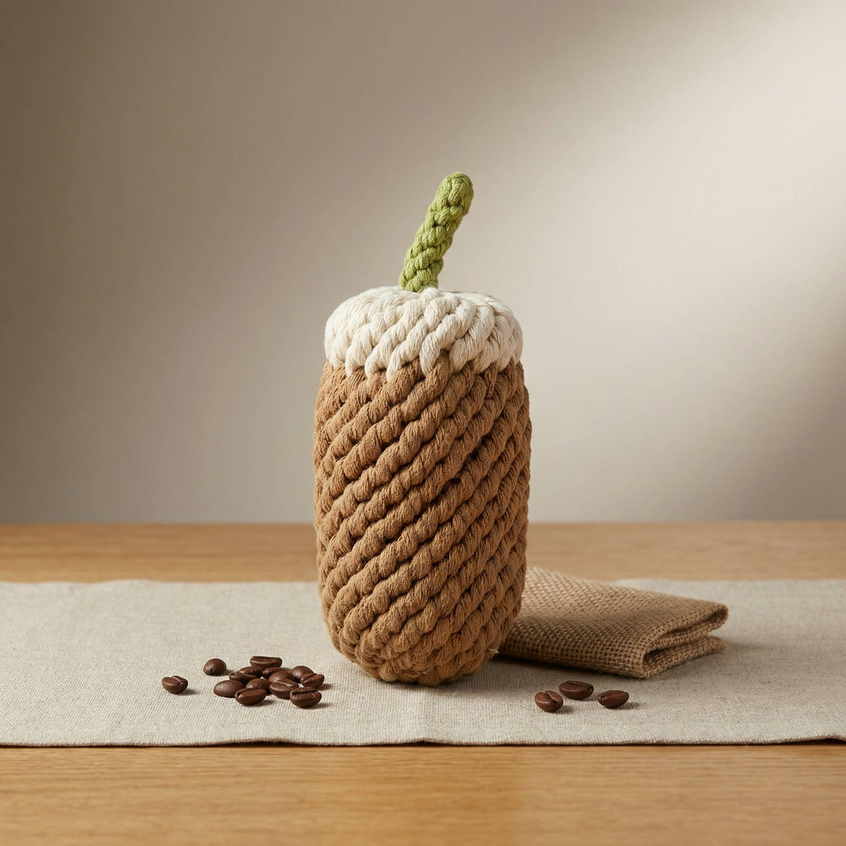 Rope toy that looks like a foamy coffee with a green straw sitting on a linen runner on a wooden table with coffee beans around it