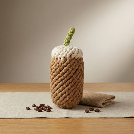 Rope toy that looks like a foamy coffee with a green straw sitting on a linen runner on a wooden table with coffee beans around it