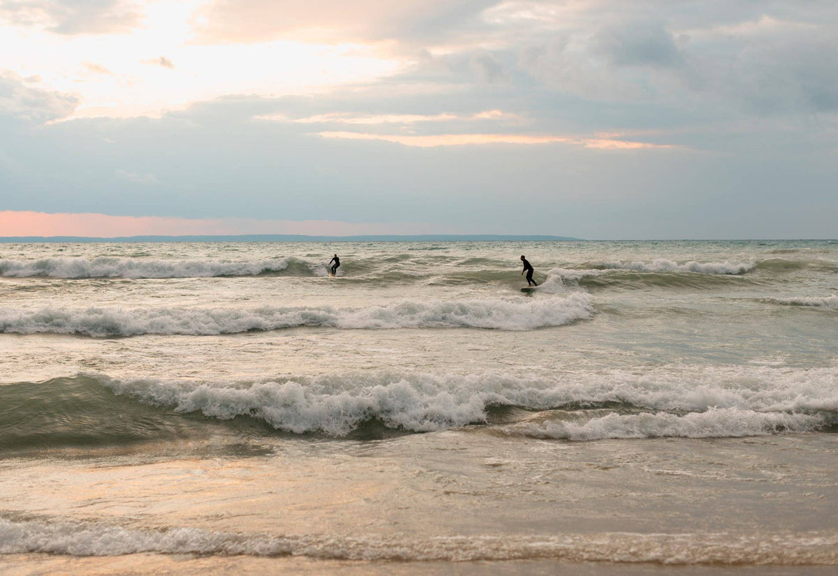 Two surfers in the ocean during sunset with waves and a cloudy sky.