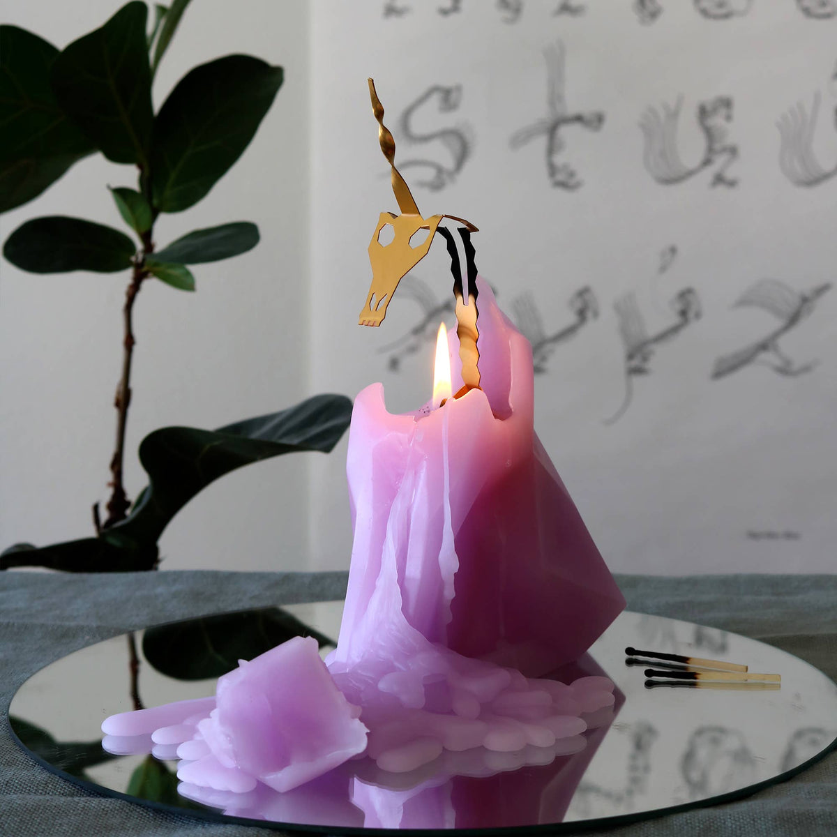 Purple candle with a unicorn-shaped wick holder on a reflective surface, with a plant and whiteboard in the background.