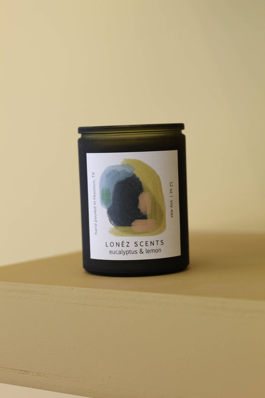 A dark amber glass candle jar with a black lid and a white label reading 'LONÉZ SCENTS eucalyptus & lemon', set on a beige wooden surface against a matching beige background.