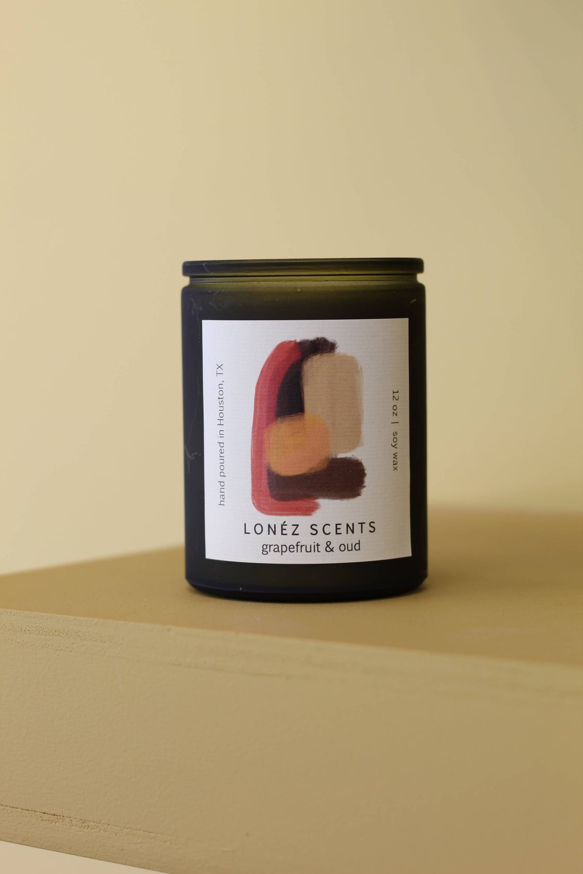 A dark glass jar candle with a black lid and a white label featuring an abstract red, beige, and orange paint stroke design. The label reads 'LONÉZ SCENTS' and 'grapefruit & oud'.