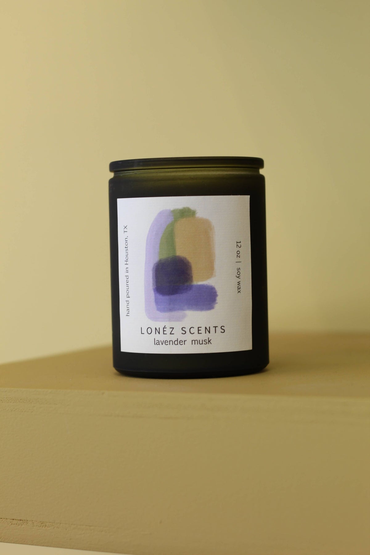 A dark glass jar candle with a black lid and a white label featuring abstract purple and green brushstrokes, labeled 'LONÉZ SCENTS lavender musk', set against a solid beige background.
