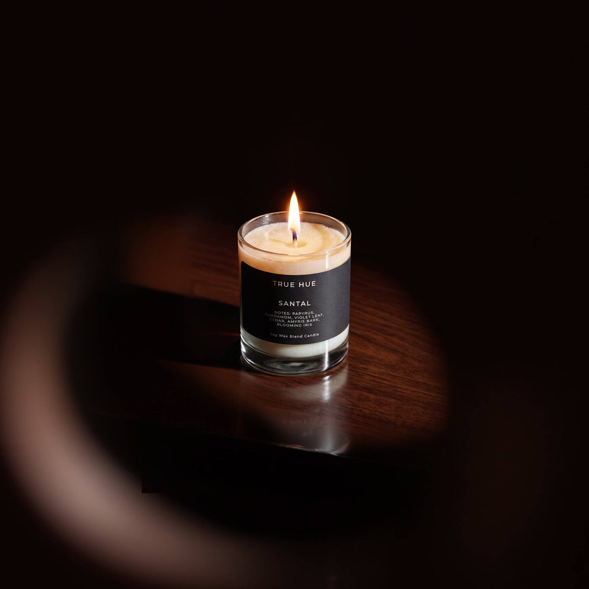 A lit candle in a clear glass jar with a black label reading TRUE HUE and SANTAL, placed on a dark wooden surface against a very dark background.