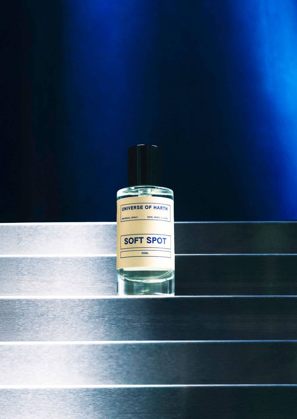 A bottle of UNIVERSE OF MANKIND fragrance named HUMAN, featuring a cream-colored label and a black cap, placed on a metallic silver staircase against a dark blue background.