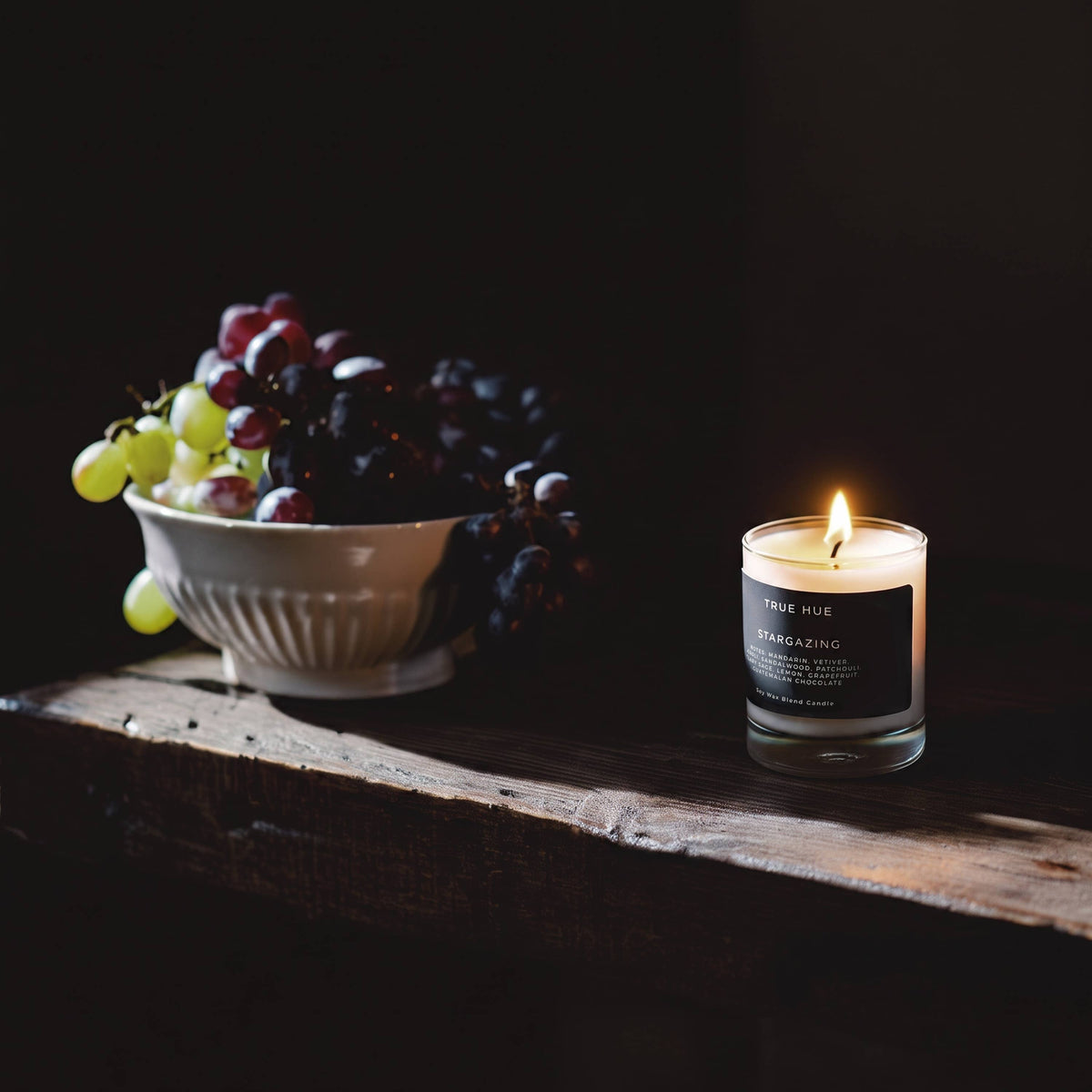 A lit scented candle in a clear glass jar sits on a rustic wooden surface next to a white bowl filled with green and purple grapes against a dark background.