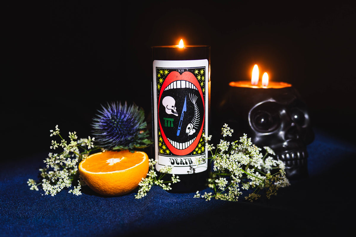 A black cylindrical candle with a gothic tarot-style 'Death' illustration, lit and standing on a dark blue surface. Surrounding it are a halved orange, white flowers, a blue thistle, and a black skull-shaped candle holder with two lit candles against a black background.