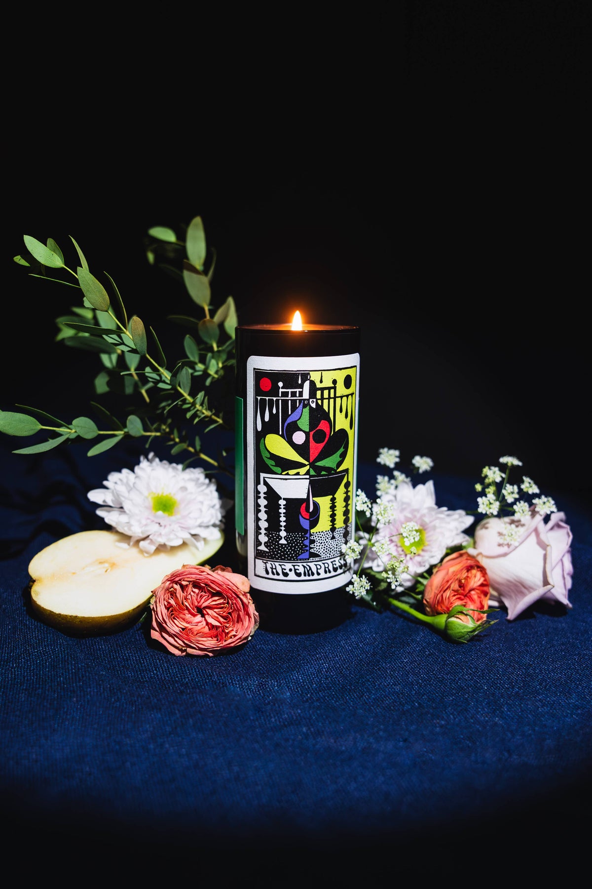 A lit black pillar candle with a colorful tarot card illustration on the front, styled on a dark blue fabric surface with flowers, greenery, and a slice of apple against a black background.