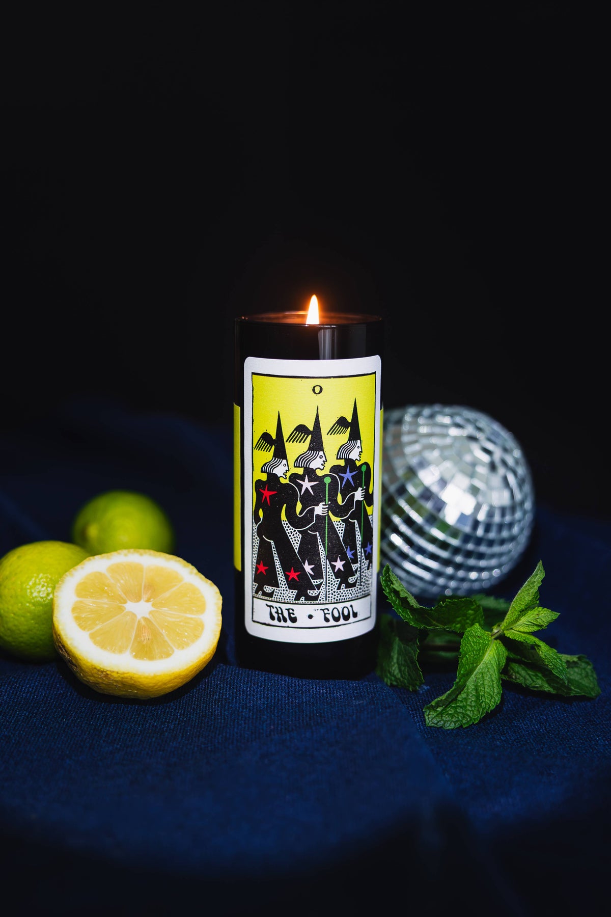 A black cylindrical candle with a lit flame and a tarot card label reading 'The Fool' is styled on a dark blue fabric surface with limes, mint leaves, and a silver disco ball against a black background.