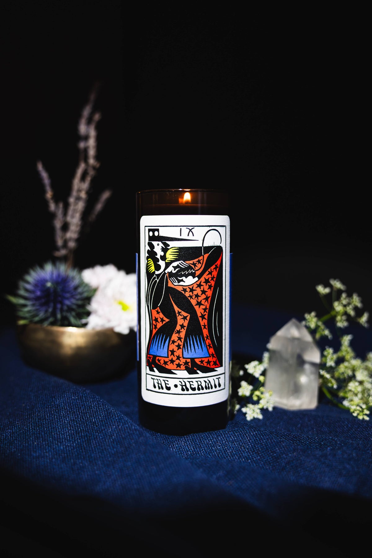 A lit black jar candle with a white label featuring the 'The Hermit' tarot card illustration, styled on a dark blue fabric surface with decorative plants and crystals against a black background.