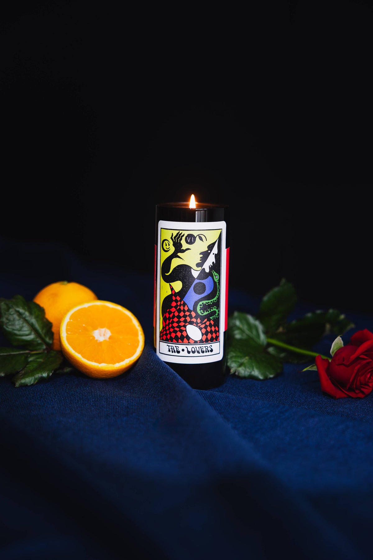 A black cylindrical candle with a colorful tarot card illustration on the front, lit with a small flame, set on a dark blue fabric surface with orange slices, green leaves, and a red rose around it against a black background.
