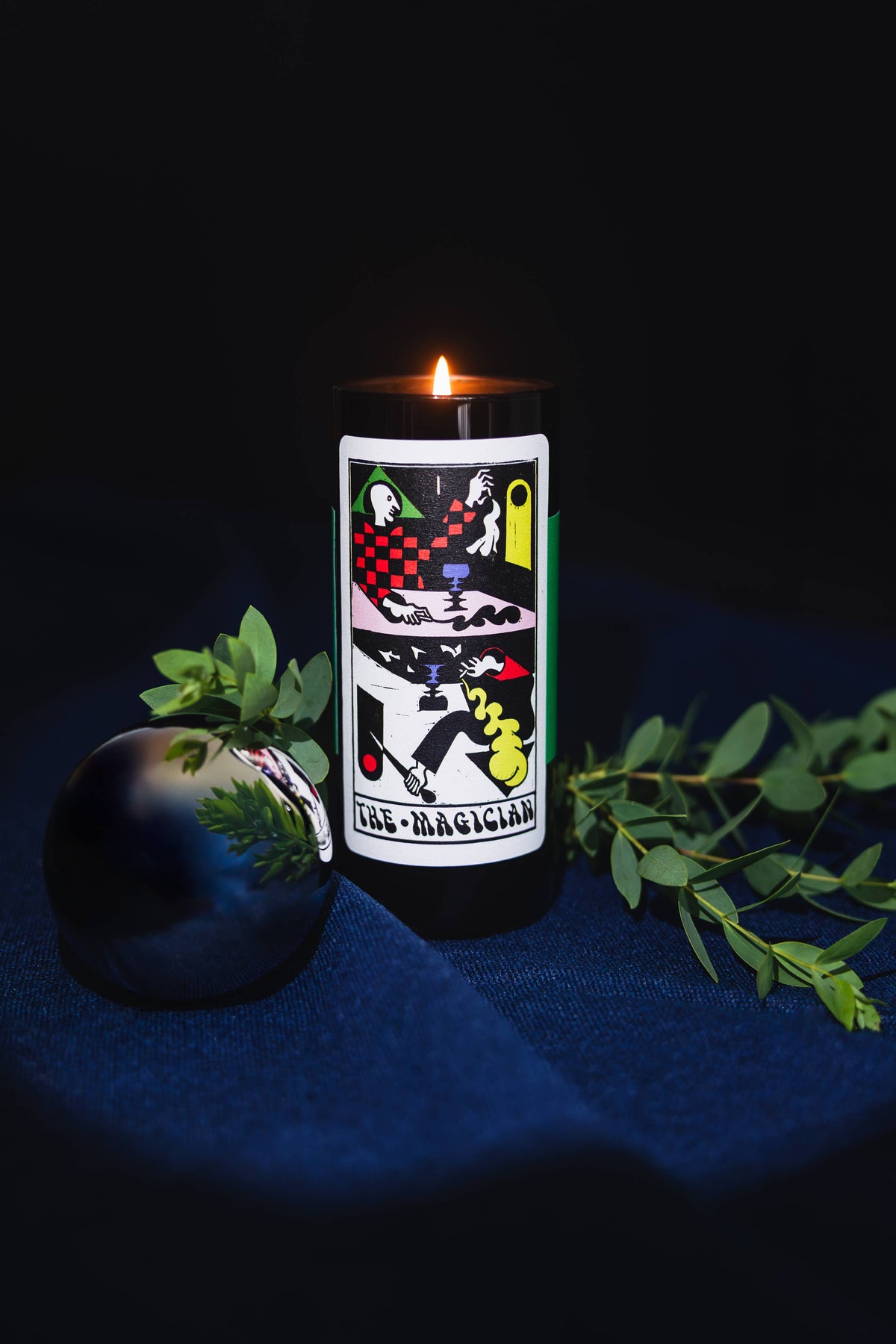 A black pillar candle with a tarot card design reading 'El Mago' and a small flame burning at the top, styled on a dark blue fabric surface with green leaves and a reflective black orb against a black background.