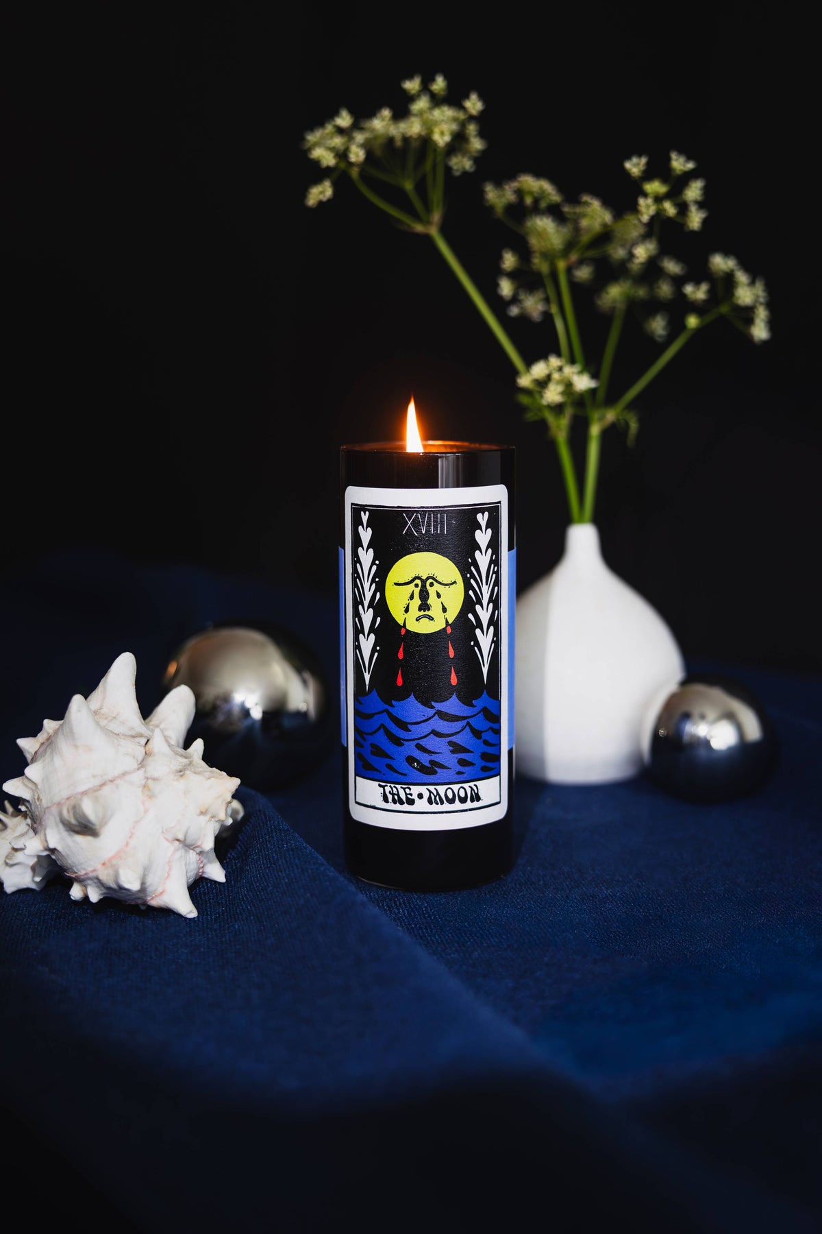 A black glass candle with a tarot card label reading 'Full Moon' is lit on a dark blue fabric surface, styled with a white seashell, a white vase with small white flowers, and two metallic spheres against a black background.