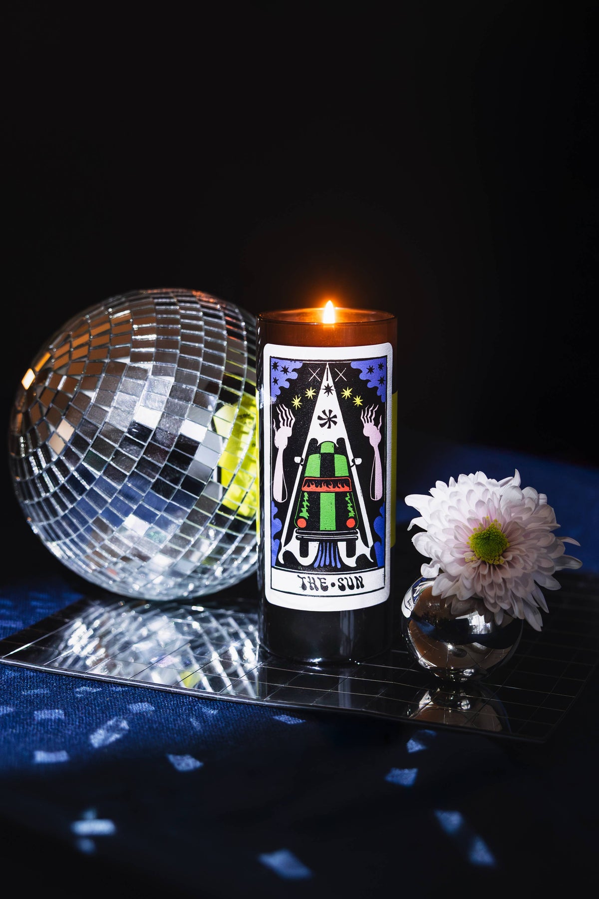 A lit black pillar candle with a colorful tarot card design on the front, styled on a reflective tray next to a disco ball and a white flower against a dark background.