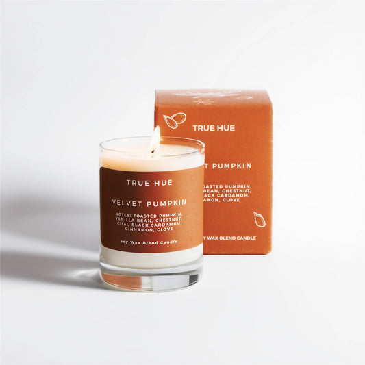 A lit True Hue Velvet Pumpkin soy wax blend candle in a clear glass jar with a brown label, standing in front of its matching brown product box on a clean white background.