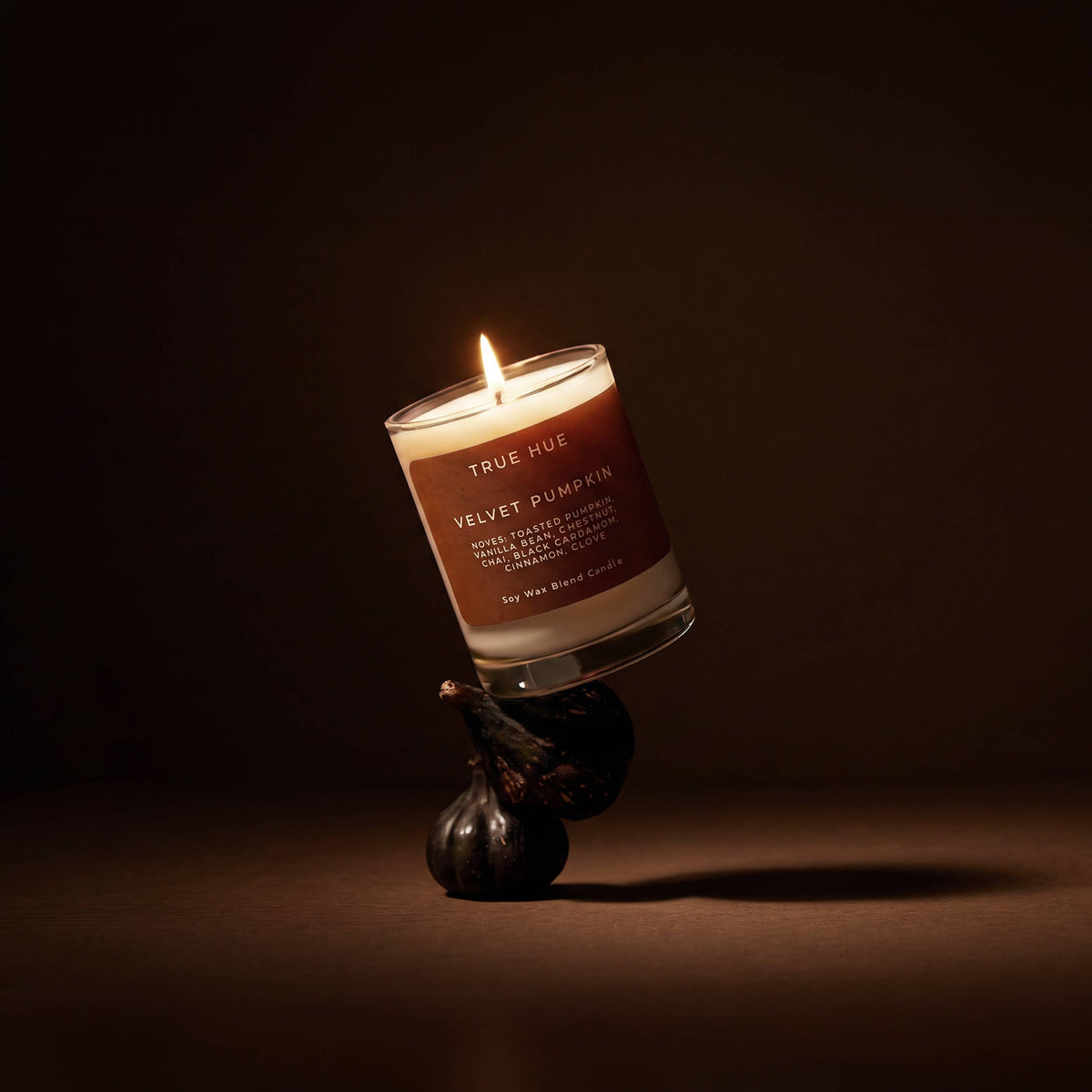 A lit candle in a clear glass vessel with a warm brown label reading TRUE HUE VELVET PUMPKIN, balanced on top of two dark figs against a dark brown background.