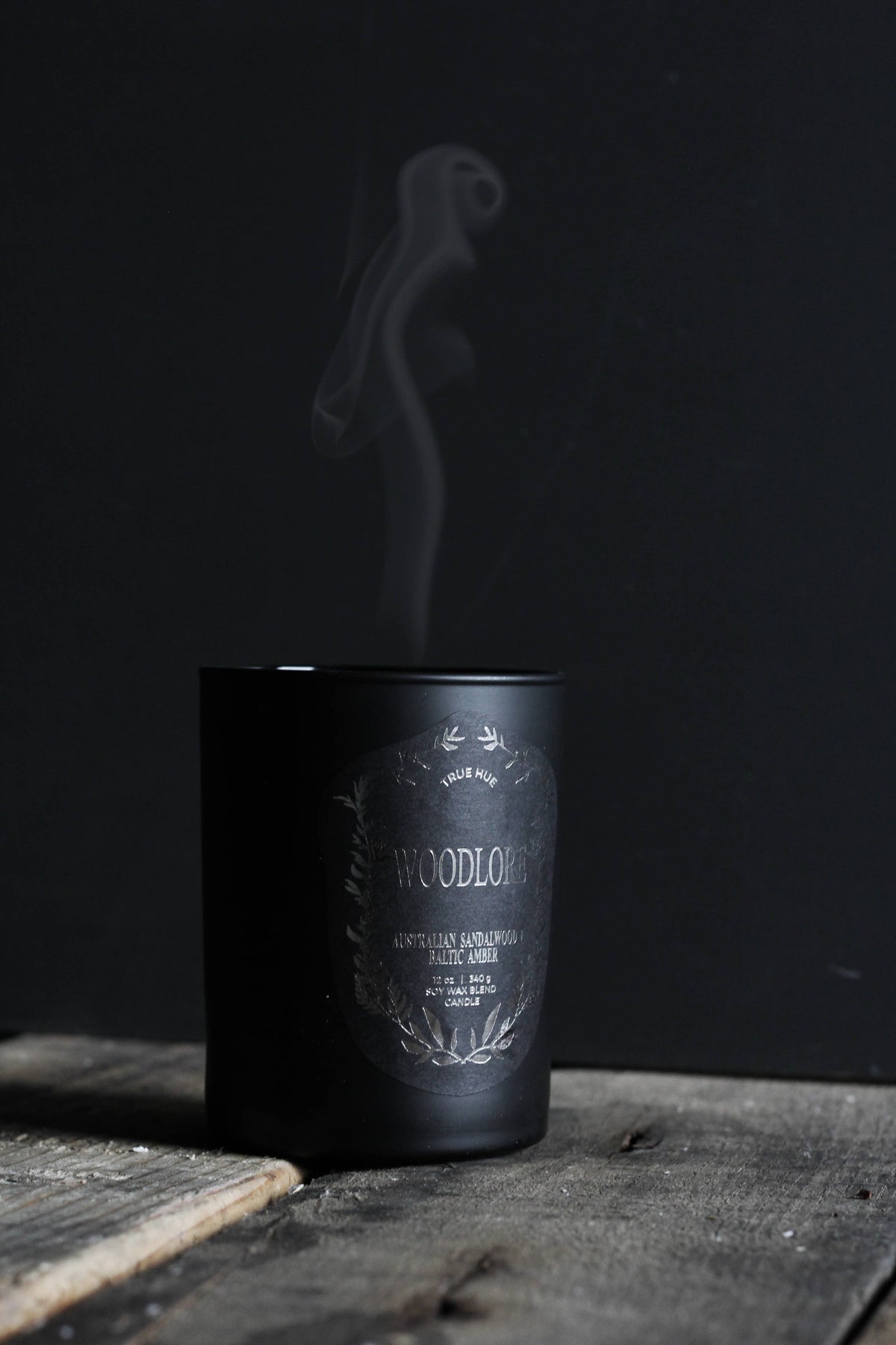 A matte black candle jar with a white decorative label reading 'WOODLAND' and 'Cedarwood & Pinewood Scents', shown on a rustic wooden surface with smoke rising from the top against a dark background.
